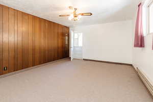 Carpeted spare room featuring wooden walls, a baseboard heating unit, and ceiling fan