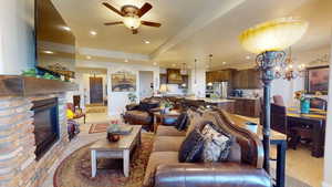 Living room with ceiling fan with notable chandelier, a stone fireplace, a tray ceiling, and light tile patterned floors