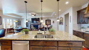 Kitchen with light stone counters, sink, an island with sink, a brick fireplace, and a raised ceiling