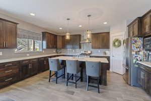 Kitchen featuring an island with sink, hanging light fixtures, wall chimney range hood, appliances with stainless steel finishes, and dark brown cabinetry