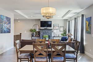 Dining area with light wood-type flooring, a stone fireplace, a tray ceiling, and a chandelier