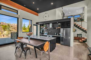 Interior space with wall chimney range hood, a high ceiling, stainless steel refrigerator with ice dispenser, an island with sink, and decorative backsplash