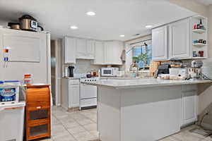 Kitchen with kitchen peninsula, white appliances, backsplash, white cabinetry, and ventilation hood