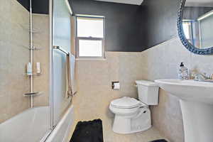 Bathroom with tile walls, tile patterned flooring, bath / shower combo with glass door, and toilet