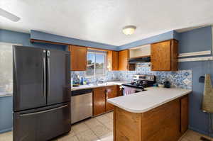 Kitchen featuring light tile patterned floors, kitchen peninsula, exhaust hood, appliances with stainless steel finishes, and decorative backsplash