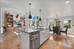 Kitchen featuring light wood-type flooring, white cabinets, hanging light fixtures, a center island with sink, and stainless steel dishwasher