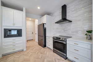 Kitchen with electric range, white cabinetry, stainless steel refrigerator, wall chimney exhaust hood, and decorative backsplash