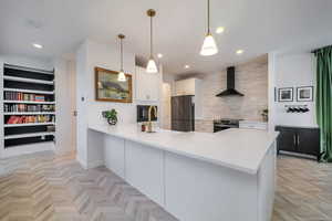 Kitchen featuring hanging light fixtures, white cabinetry, kitchen peninsula, stainless steel appliances, and wall chimney range hood