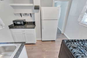 Kitchen with dark stone counters, light wood-type flooring, white refrigerator, range, and white cabinetry