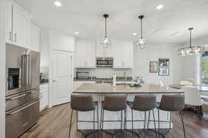 Kitchen with an island with sink, decorative light fixtures, dark wood-type flooring, white cabinetry, and appliances with stainless steel finishes