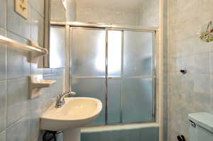 Full bathroom with tile walls, toilet, sink, and combined bath / shower with glass door