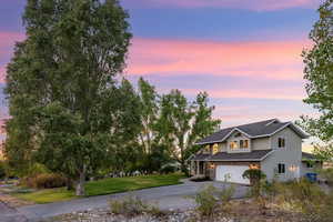 View of front of home with mature shade trees, a yard and a garage at sunset