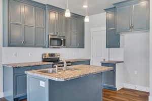 Kitchen with dark wood-type flooring, stainless steel appliances, backsplash, a center island with sink, and decorative light fixtures