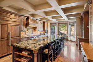 Kitchen featuring dark stone counters, wood-type flooring, a large island with sink, coffered ceiling, and decorative backsplash