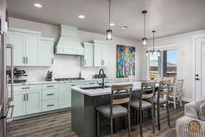 Kitchen featuring dark wood-type flooring, custom exhaust hood, decorative light fixtures, stainless steel gas stovetop, and a kitchen island with sink