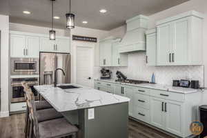 Kitchen featuring sink, stainless steel appliances, a center island with sink, light stone countertops, and premium range hood