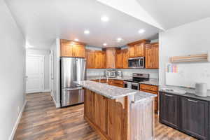 Kitchen with a breakfast bar, light stone counters, stainless steel appliances, a center island with sink, and dark hardwood / wood-style flooring