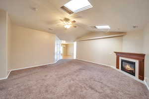 Unfurnished living room with a tiled fireplace, ceiling fan, light colored carpet, and lofted ceiling with skylight