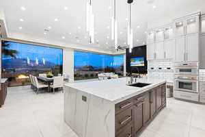 Kitchen with light stone counters, stainless steel double oven, sink, white cabinetry, and an island with sink