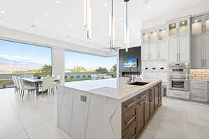 Kitchen with sink, light stone counters, double oven, an island with sink, and decorative light fixtures