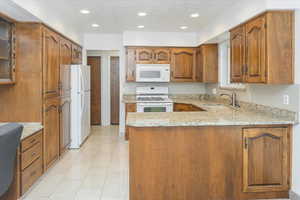 Kitchen with light tile patterned flooring, sink, kitchen peninsula, white appliances, and light stone countertops