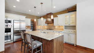 Kitchen featuring a center island with sink, glass insert cabinets, built in appliances, white cabinetry, and pendant lighting