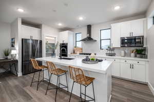 Kitchen with appliances with stainless steel finishes, wall chimney exhaust hood, a kitchen island with sink, and white cabinetry