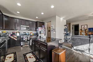 Kitchen with dark brown cabinets, dark wood-type flooring, a kitchen island, appliances with stainless steel finishes, and dark stone countertops