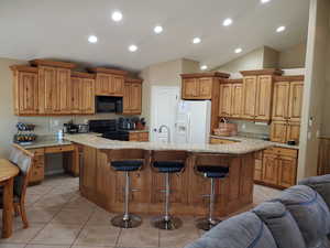 Kitchen featuring light stone counters, an island with sink, vaulted ceiling, black appliances, and a kitchen bar