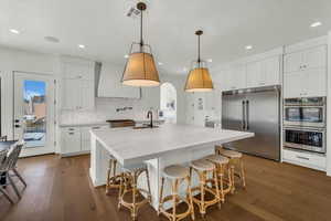 Kitchen with a kitchen island with sink, white cabinetry, hanging light fixtures, custom exhaust hood, and appliances with stainless steel finishes