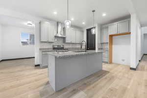 Kitchen featuring light hardwood / wood-style floors, an island with sink, wall chimney exhaust hood, decorative light fixtures, and gas stove