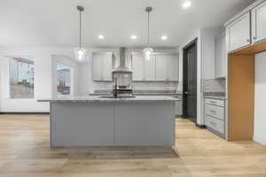 Kitchen featuring an island with sink, decorative light fixtures, wall chimney exhaust hood, light hardwood / wood-style flooring, and light stone countertops