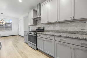 Kitchen with gray cabinetry, light stone counters, light hardwood / wood-style flooring, gas stove, and wall chimney range hood