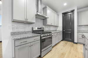 Kitchen with light hardwood / wood-style flooring, wall chimney range hood, gray cabinets, gas stove, and decorative backsplash