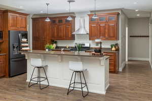 Kitchen featuring a center island with sink, wall chimney exhaust hood, stainless steel appliances, a breakfast bar area, and dark hardwood / wood-style flooring