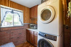 Clothes washing area featuring stacked washing maching and dryer, brick wall, wood walls, cabinets, and dark hardwood / wood-style floors