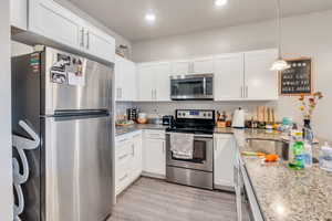 Kitchen with hanging light fixtures, white cabinetry, light stone counters, stainless steel appliances, and light wood-type flooring