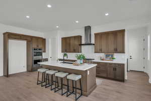 Kitchen with stainless steel double oven, light hardwood / wood-style flooring, a kitchen island with sink, and wall chimney exhaust hood