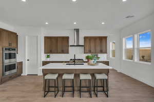 Kitchen with a kitchen island with sink, light hardwood / wood-style flooring, wall chimney exhaust hood, and stainless steel double oven