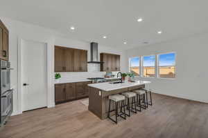 Kitchen with light wood-type flooring, a breakfast bar, sink, wall chimney range hood, and a center island with sink