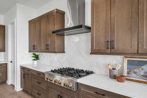 Kitchen featuring decorative backsplash, light wood-type flooring, dark brown cabinets, stainless steel gas cooktop, and wall chimney range hood