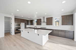 Kitchen featuring sink, wall chimney exhaust hood, stainless steel appliances, light hardwood / wood-style flooring, and an island with sink