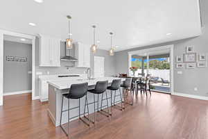 Kitchen with a kitchen island with sink, white cabinets, dark wood-type flooring, wall chimney range hood, and hanging light fixtures