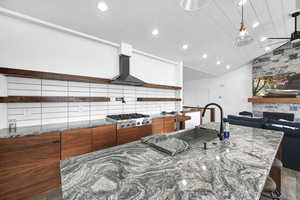 Kitchen featuring wall chimney range hood, stainless steel gas cooktop, dark stone counters, sink, and a spacious island