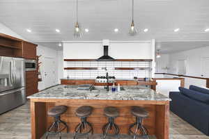 Kitchen with light stone counters, a kitchen island with sink, hardwood / wood-style flooring, appliances with stainless steel finishes, and range hood