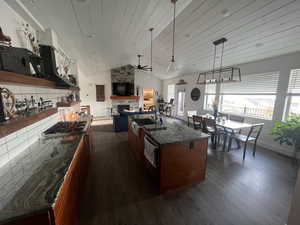 Kitchen featuring ceiling fan, hanging light fixtures, a stone fireplace, a kitchen island with sink, and dark hardwood / wood-style flooring