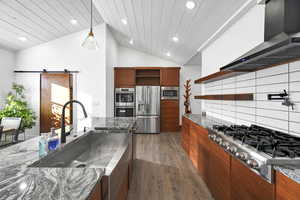 Kitchen featuring lofted ceiling, sink, wall chimney range hood, a barn door, and appliances with stainless steel finishes