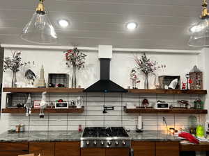 Kitchen featuring dark stone countertops, wall chimney range hood, stainless steel gas stovetop, and backsplash