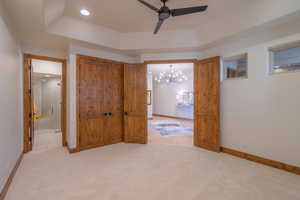 Unfurnished bedroom featuring ceiling fan with notable chandelier, light colored carpet, a raised ceiling, and connected bathroom