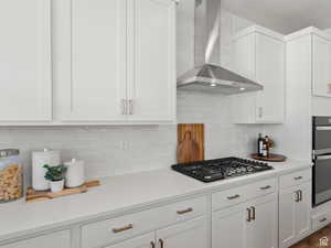 Kitchen with wall chimney exhaust hood, backsplash, stainless steel appliances, and white cabinetry